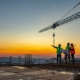 3 construction workers on top of a building being constructed with a crane in the background at sunset.